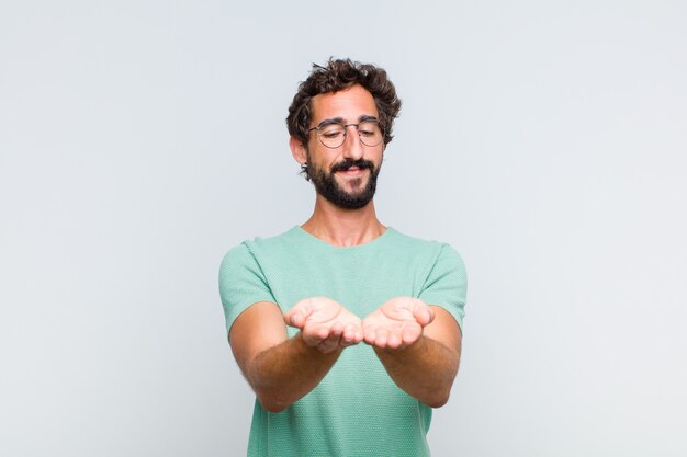 Joven barbudo sonriendo felizmente con mirada amable, confiada y positiva, ofreciendo y mostrando