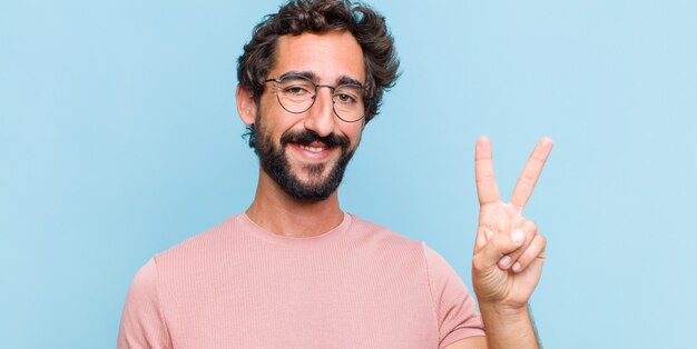 Joven barbudo sonriendo y mirando amigable, mostrando el número dos o el segundo con la mano hacia adelante, contando hacia atrás