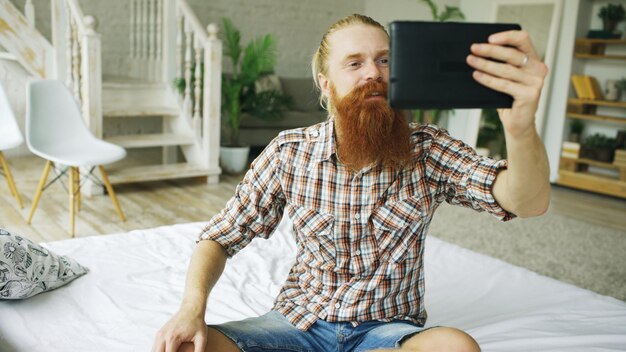 Joven barbudo usando una tableta con video chat sentado en la cama en casa