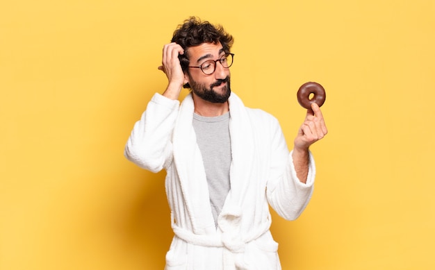 Joven barbudo vestido con albornoz con una dona de chocolate