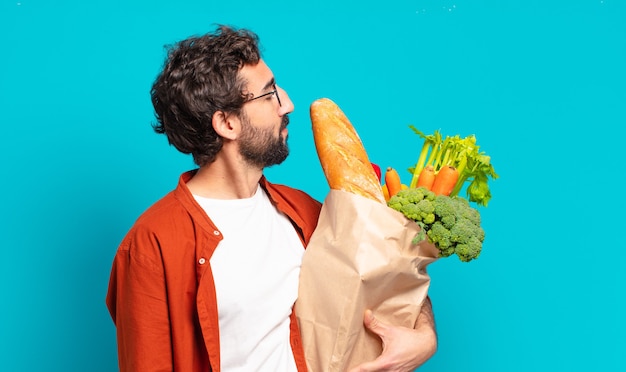 Joven barbudo en vista de perfil mirando para copiar espacio adelante, pensando, imaginando o soñando despierto y sosteniendo una bolsa de verduras