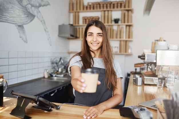 Joven barista entregando una taza de capuchino o tarde al cliente mirando a la cámara sonriendo Preparando café recién hecho para un gran sabor