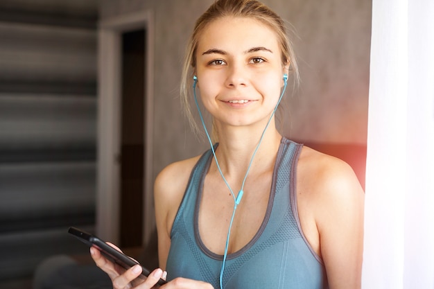 Joven bastante deportiva en casa escucha música en auriculares desde el teléfono inteligente. Día soleado