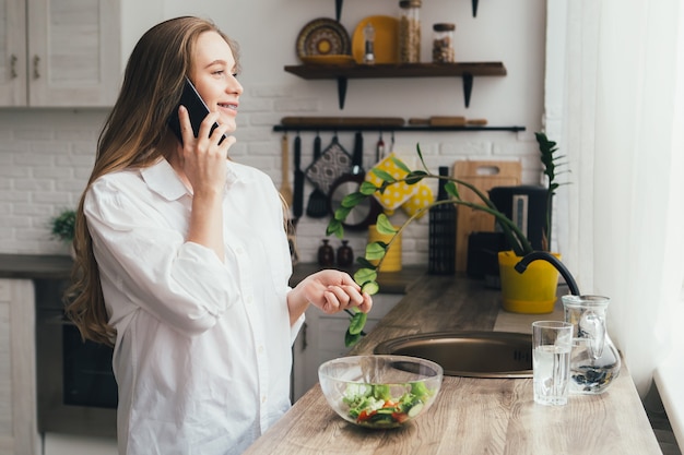 Joven bastante embarazada está hablando por teléfono en la cocina y masticando un pepino de una ensalada