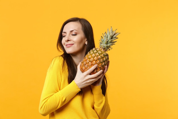 Una joven bastante sonriente con ropa informal que mantiene los ojos cerrados sostiene fruta fresca de piña madura aislada en un fondo amarillo anaranjado. El estilo de vida vívido de la gente relaja el concepto de vacaciones. Simulacros de espacio de copia