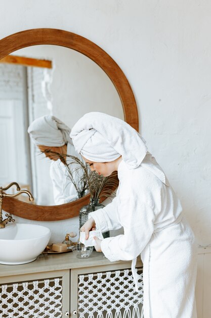 Una joven en bata de baño con una toalla envuelta alrededor de su cabeza está extendiendo una mascarilla orgánica en un tazón en el baño.