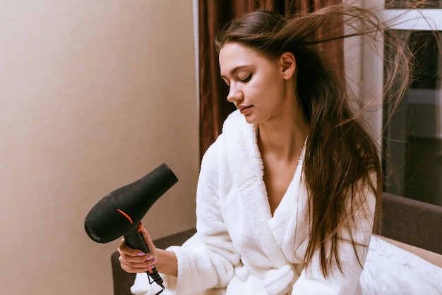 Una joven con una bata de felpa blanca se seca el cabello con un secador de pelo