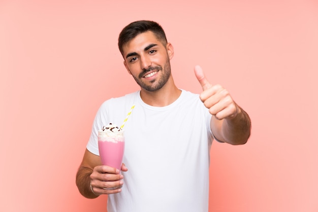 Joven con batido de fresa sobre pared rosa aislado con los pulgares hacia arriba porque algo bueno