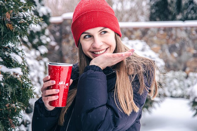 Una joven bebe una bebida caliente de una taza térmica roja en invierno