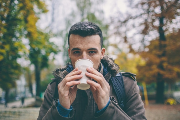 Joven beber café o té para llevar fuera en el parque o en la calle. Mira directamente a la cámara y posa. Otoño. Está frío afuera.