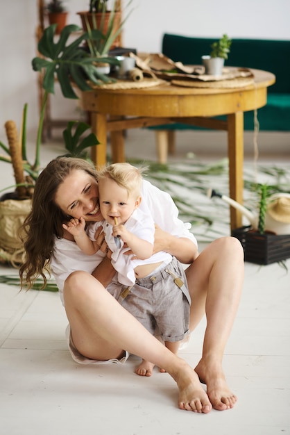 Una joven y bella mamá jugando con su bebé en el piso junto a las plantas y el sofá verde