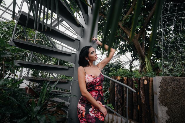 Una joven y bella morena posa cerca de una escalera de caracol en el jardín botánico entre los densos matorrales del bosque tropical. Spa.