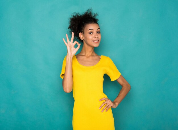 Joven y bella mujer afroamericana mostrando signo ok. Chica alegre gesticulando que todo es genial, fondo de estudio azul