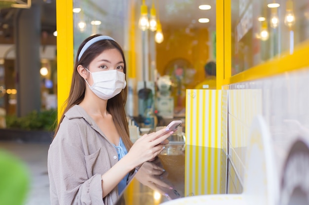 Joven y bella mujer asiática con una máscara para prevenir enfermedades está sentada y esperando una bebida frente a una cafetería. mientras sostiene un teléfono inteligente y mira a la cámara.