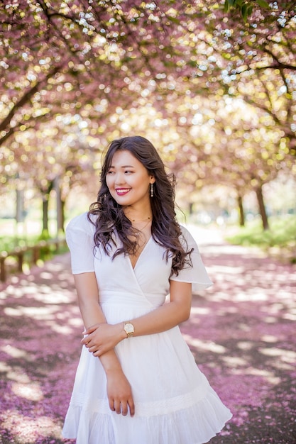 Una joven y bella mujer asiática con un vestido blanco camina en un parque de flores. Sakura. Árboles florecientes. Primavera.