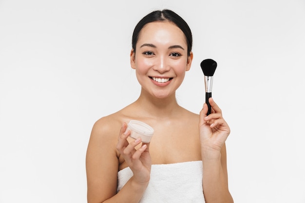 Joven y bella mujer bastante asiática con piel sana posando desnuda aislada sobre pared blanca con pincel de maquillaje y polvo.