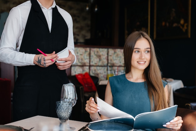 Una joven y bella mujer en un buen restaurante mira el menú y hace un pedido a un joven camarero con un elegante delantal. Servicio al Cliente.