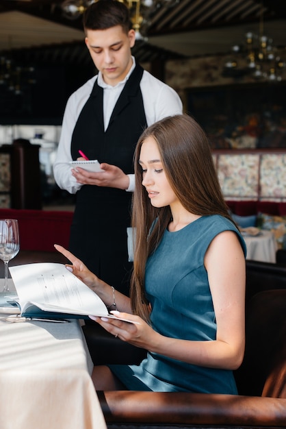 Una joven y bella mujer en un buen restaurante mira el menú y hace un pedido a un joven camarero con un elegante delantal. Servicio al Cliente.