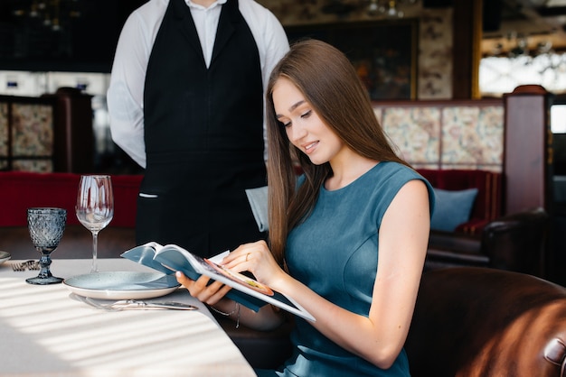 Una joven y bella mujer en un buen restaurante mira el menú y hace un pedido a un joven camarero con un elegante delantal. Servicio al Cliente.