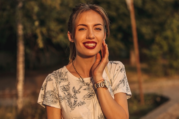 joven y bella mujer en camiseta blanca y pantalones cortos de mezclilla con abrigo rojo se encuentra al lado del grueso árbol en la superficie de verano