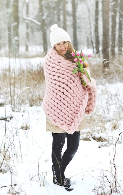 Joven y bella mujer caucásica en ropa de invierno y una manta gigante de tejer rosa pastel con flores de primavera caminando en el bosque nevado soñando con un sombrero blanco de primavera