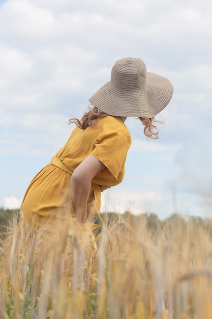 Una joven y bella mujer embarazada con un vestido amarillo y un sombrero camina a través de un campo de trigo naranja en un día soleado de verano