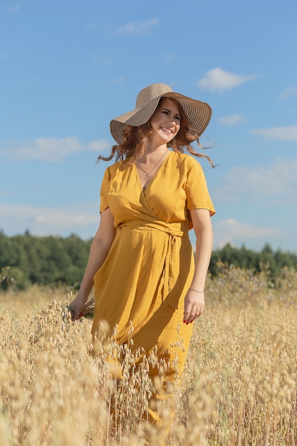 Una joven y bella mujer embarazada con un vestido amarillo y un sombrero camina a través de un campo de trigo naranja en un día soleado de verano