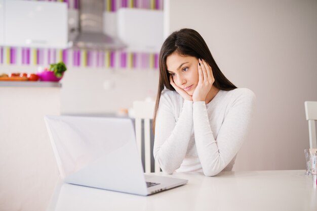 Joven y bella mujer enfocada está leyendo cuidadosamente cosas en su computadora portátil mientras está sentado a la mesa en una habitación muy luminosa.