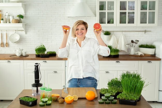 Una joven y bella mujer está cortando frutas y riéndose en la mesa de la cocina por un batido de vitaminas hecho de microverduras y frutas.