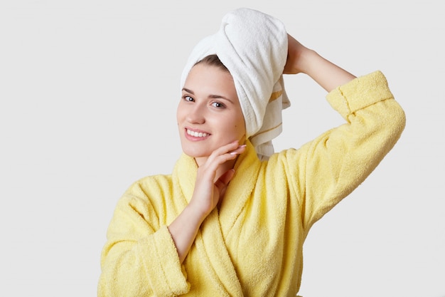 La joven y bella mujer europea tiene una toalla en la cabeza, usa una bata de baño, una sonrisa encantadora, una piel suave y saludable