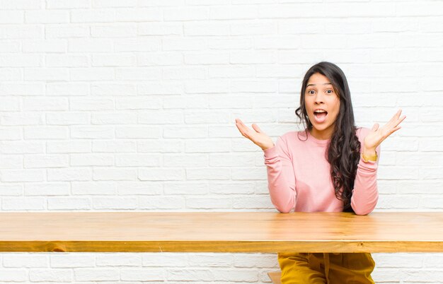 Joven y bella mujer latina se siente conmocionada y emocionada, riendo, asombrada y feliz por una sorpresa inesperada sentado frente a una mesa