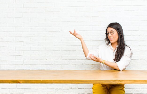 Joven y bella mujer latina sonriendo con orgullo y confianza, sintiéndose feliz y satisfecha y mostrando un copyspace sentado frente a una mesa