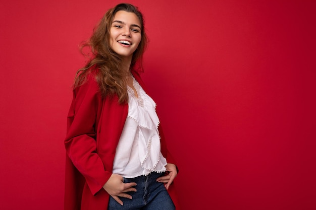 Joven y bella mujer morena con estilo europeo vistiendo blusa blanca y rebeca rojo aislado sobre fondo rojo con emociones sinceras positivas. Mirada simple y natural a la cámara. Espacio libre.