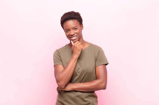 Joven y bella mujer negra sonriendo, disfrutando de la vida, sintiéndose feliz, amigable, satisfecha y despreocupada con la mano en la barbilla contra la pared rosa