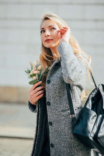 Foto la joven y bella mujer rubia con el pelo ondulado largo y rubio sonríe a la gente en la calle y sosteniendo su cabello rubio, la mochila y el pequeño ramo.