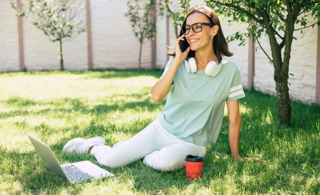 Joven y bella mujer segura feliz moderna en auriculares con un teléfono inteligente está trabajando en una computadora portátil mientras está sentado en el césped.