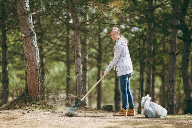 Joven y bella mujer sonriente limpiando y usando un rastrillo para la recolección de basura cerca de las bolsas de basura en el parque o el bosque. Problema de contaminación ambiental. Detener la basura de la naturaleza, el concepto de protección del medio ambiente.