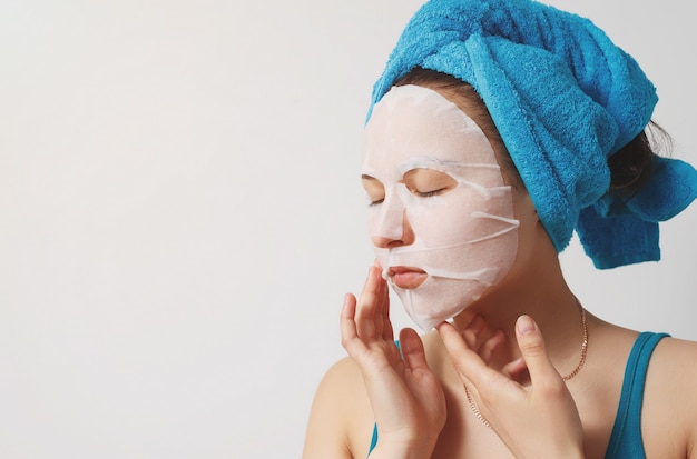Una joven y bella mujer usa una mascarilla facial de tela cosmética hidratante con una toalla envuelta alrededor de su cabeza. en la pared blanca