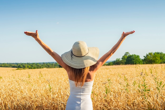 Joven y bella mujer con un vestido blanco y un sombrero levanta sus manos en el campo