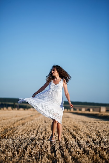 Una joven y bella mujer con un vestido blanco de verano se encuentra en un campo de trigo segado con enormes gavillas de heno, disfrutando de la naturaleza. Naturaleza en el pueblo. Enfoque selectivo