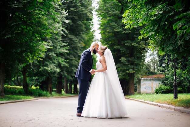 Una joven y bella novia y su esposo está de pie en un parque de verano con un ramo de flores