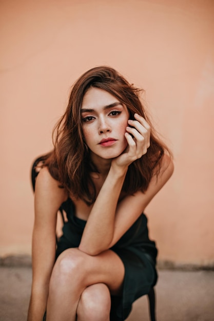 Joven bien construida con peinado ondulado marrón y maquillaje de moda con vestido oscuro posando en una silla al aire libre y mirando a la cámara contra el fondo de la pared de melocotón