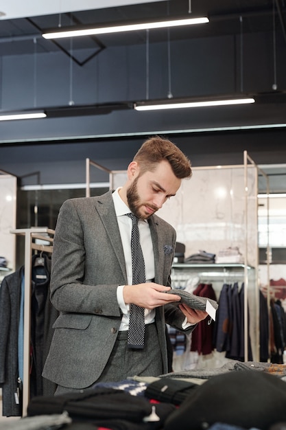 Joven bien vestido mirando a través de la nueva colección en una boutique contemporánea y eligiendo un nuevo gorro de punto