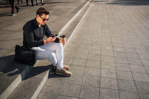 Un joven bien vestido se sienta en los escalones y mira el teléfono. Él sostiene la taza de café. La bolsa de cuero está a su lado. Está soleado afuera.