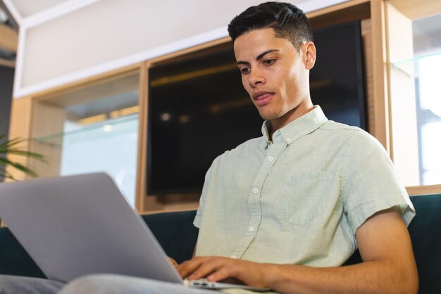 Un joven biracial trabajando en una computadora portátil sentado en un sofá en una oficina de negocios moderna