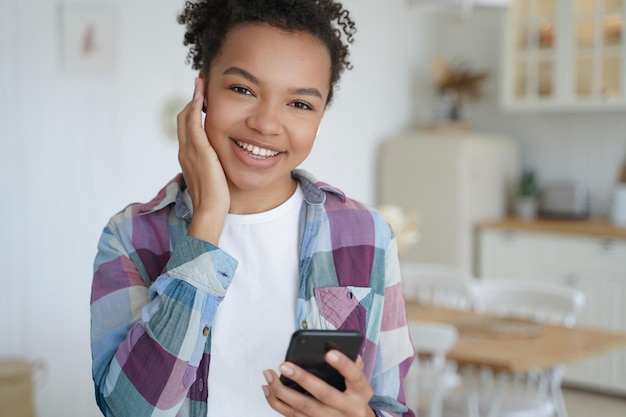 La joven birracial moderna y feliz que sostiene un teléfono inteligente escucha música con un auricular y usa aplicaciones musicales