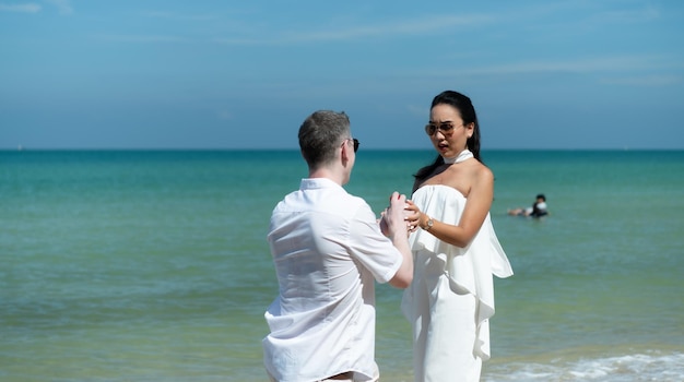 Un joven blanco pidiendo a una chica asiática que se case con un anillo de diamantes en una playa