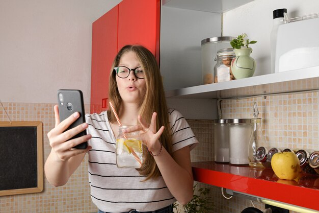 Joven blogger hablando con la cámara de un teléfono, haciendo un live en las redes sociales mostrando una bebida mientras está sentado en la mesa de la cocina