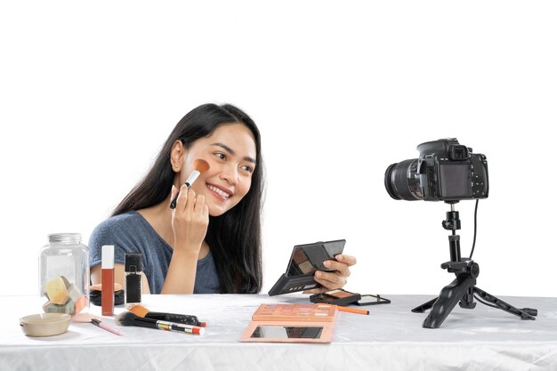 Joven blogger de video femenina sonriente con pincel mostrando el producto cosmético en la mesa a la cámara