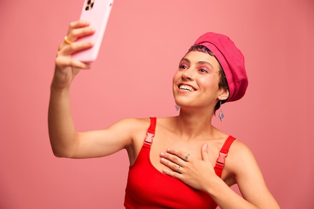 Una joven bloguera con cabello rosa teñido y un corte de pelo corto se toma una foto por teléfono y transmite una sonrisa con ropa elegante y un sombrero en un estilo monocromático de fondo rosa